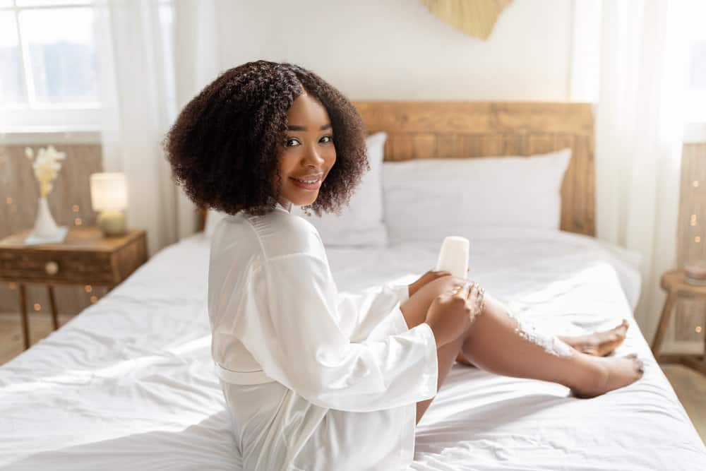 A black lady with curly head hair sitting on the bed preparing to use a razor blade to remove all the hair from her legs.