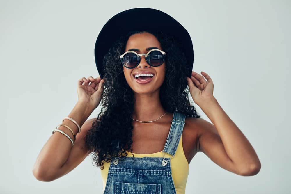 An attractive young woman with curly, fine hair wearing a yellow shirt and blue jean overalls.