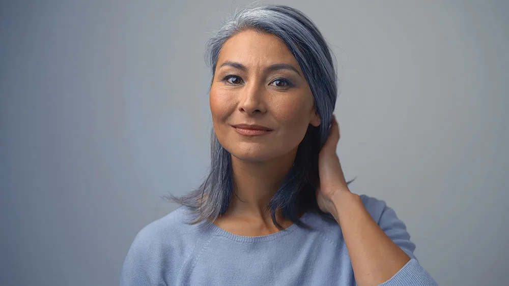 Removing Hair Color to Go Grey at Home: DIY How-to Guide 5 Elderly Asian woman with salt and pepper hair wearing a light blue sweater.