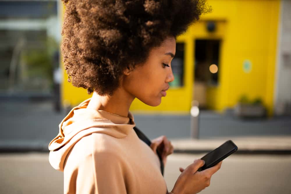Beautiful young black woman with a curly afro after drying her wet hair, reading about hair masks on her new iPhone.
