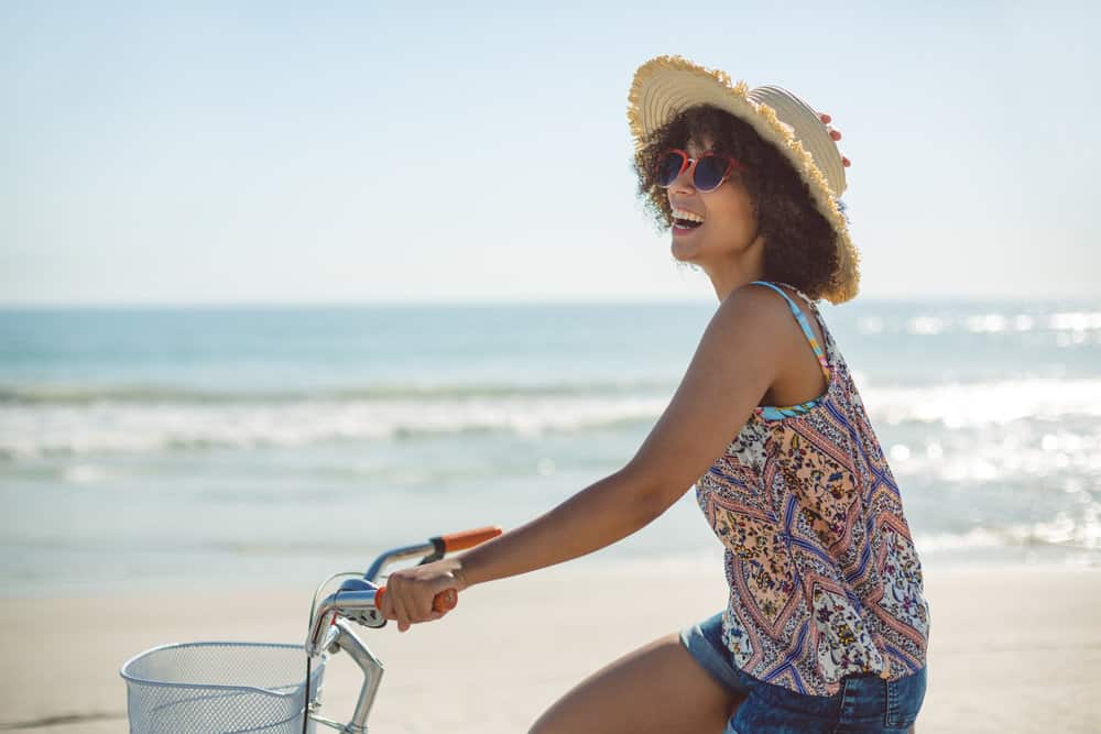 A lady with dark brown naturally wavy hair strands riding a bike on a sunny day at the beach.