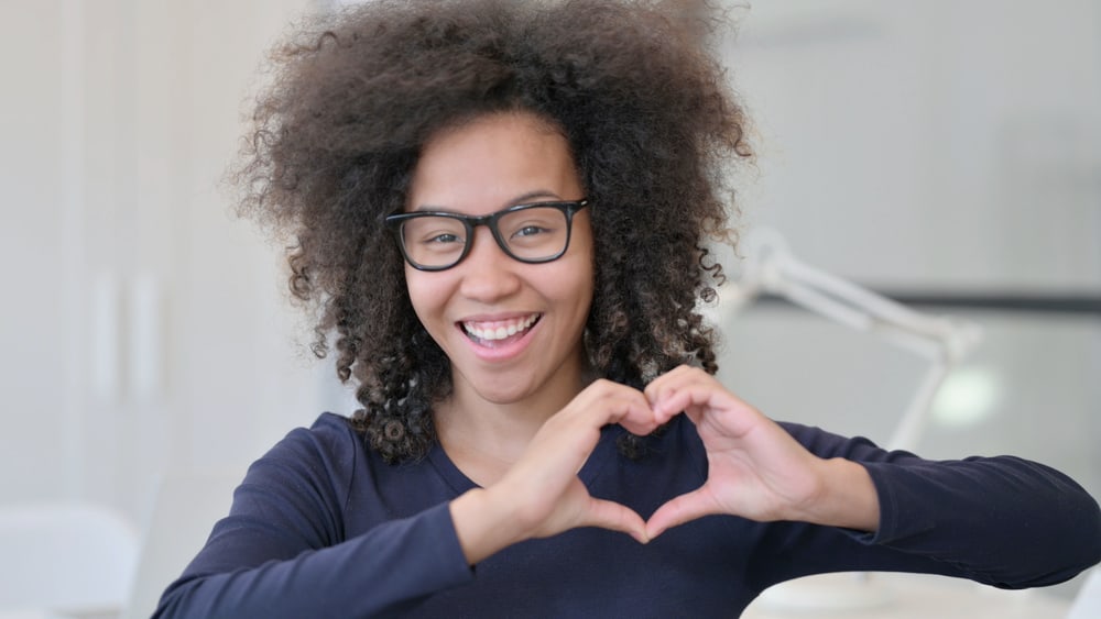 A young black female with a wolf haircut is making a heart shape with her hands while wearing a blue shirt.