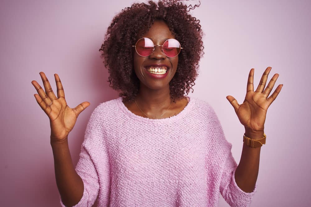 Happy young woman with dark skin wearing a pink sweater and a curly wig creating a very realistic look.
