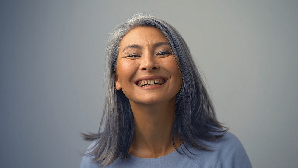 A lovely Asian woman is going through a gray hair change after a few washes with purple shampoo.