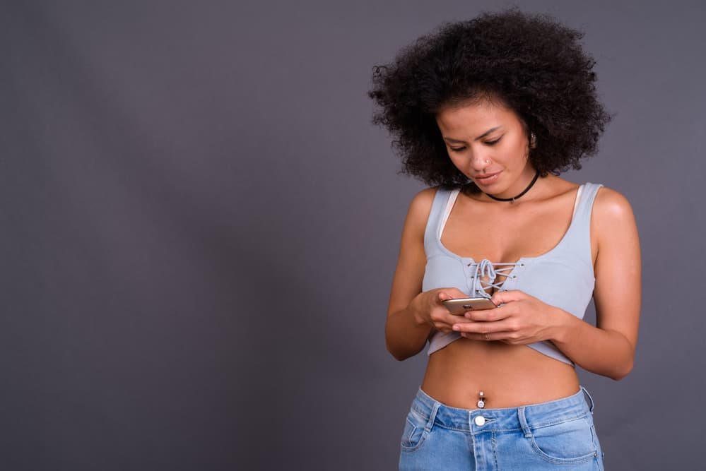A black girl with a belly ring using an iPhone to research the difference between baby shampoo and dry shampoo.