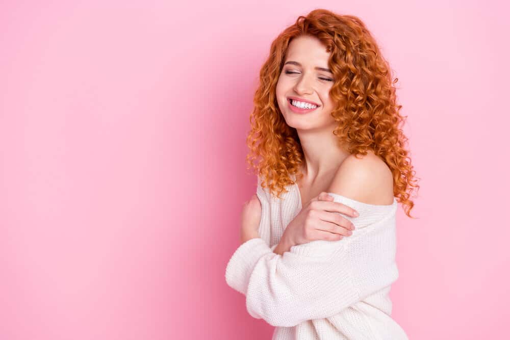 A white woman wearing an off-the-shoulder sweater with coily hair styled with Luster's Pink Oil Moisturizer Hair Lotion.