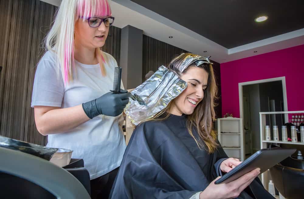 While applying hair dye to a beautiful young lady, the hairdresser is looking at a digital tablet.