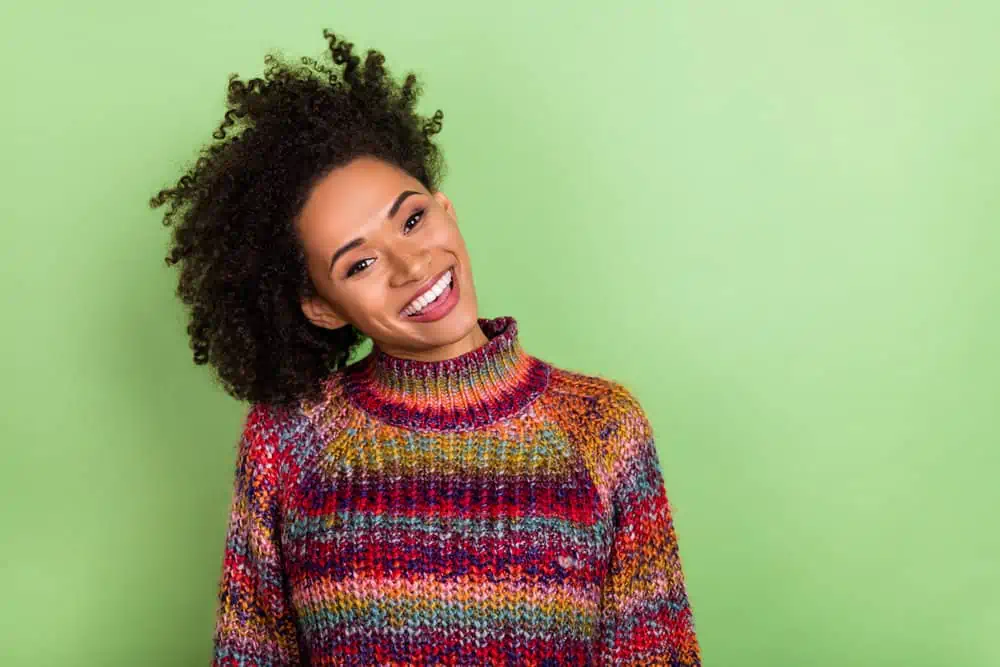 A cute young black lady wearing a colorful sweater that's experiencing hair shaft damage after using hard hair dye.