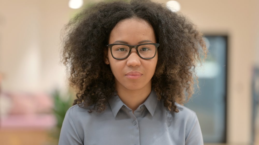 Light-skinned black girl with wavy hair wearing a shag cut with black eyeglasses and a gray dress shirt.