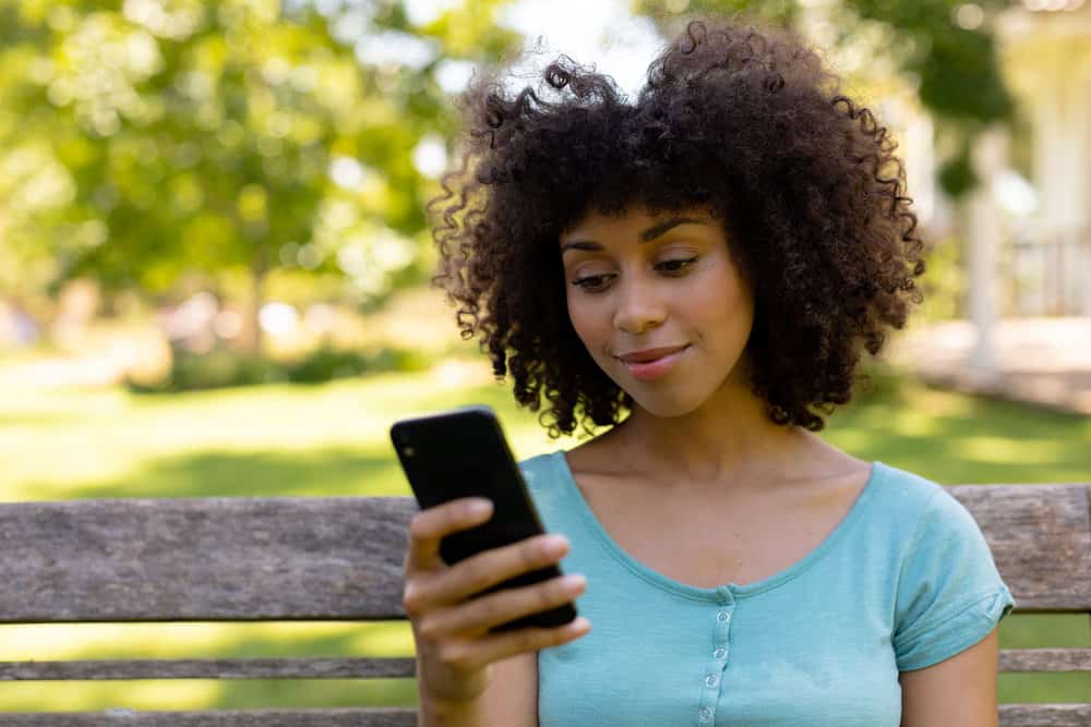 Black women wearing make-up, a blue shirt, and naturally curly hair while using an iPhone.