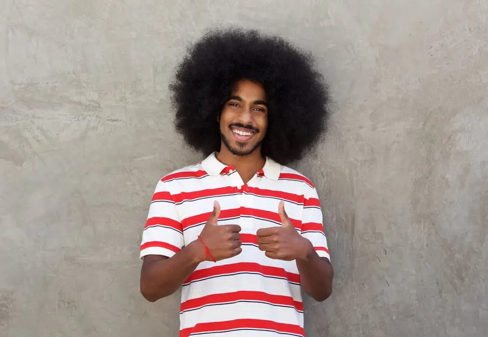 A young man with both thumbs up wearing his hair long after not getting a hair cut for several years.