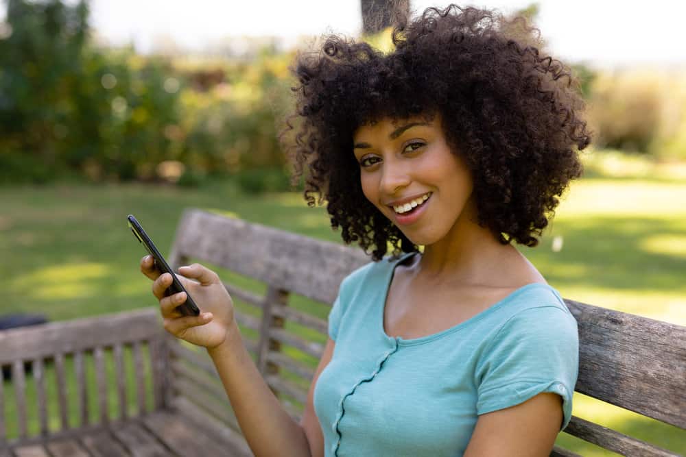 A cute black lady sitting on a park bench wearing a blue shirt using a mobile phone.