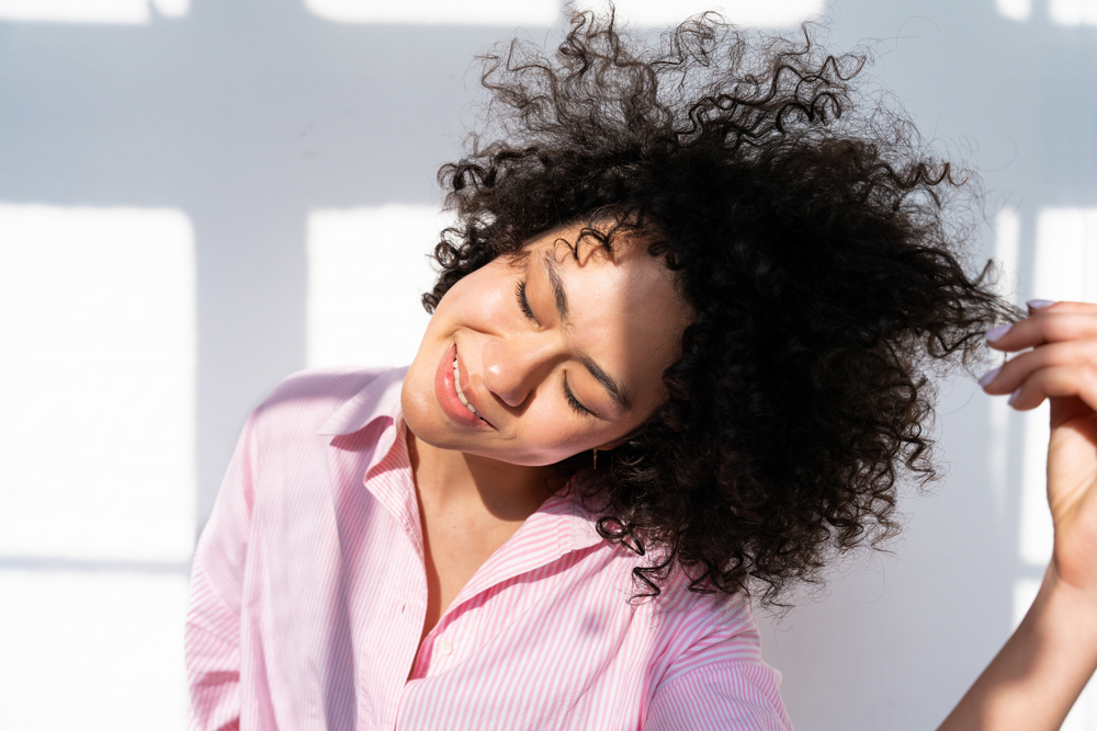 A lady with flawless skin and thick hair running her fingers through her curls and admiring her hair's health.