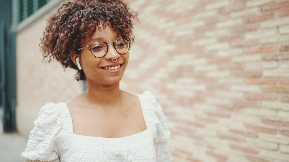 Young woman with really curly hair walking outdoors while listening to a podcast about hair loss using AirPods.