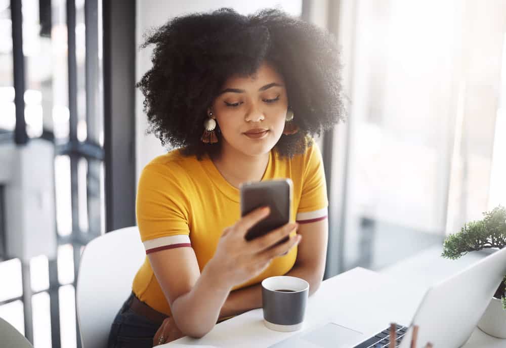 A beautiful dark-skinned woman with natural curls wearing a gold t-shirt researching blond hair on her iPhone.