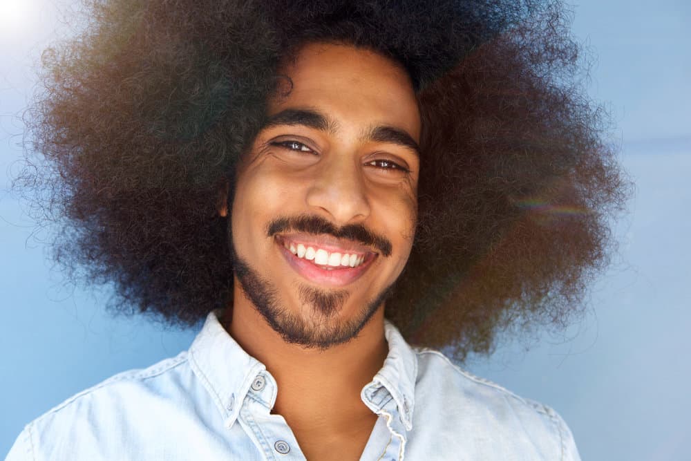 A smiling young man with facial hair and a full head with shoulder-length curls in their natural texture.