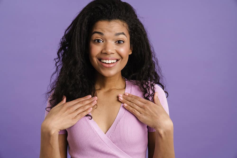 Young black woman smiling while holding hands on her chest isolated over purple background