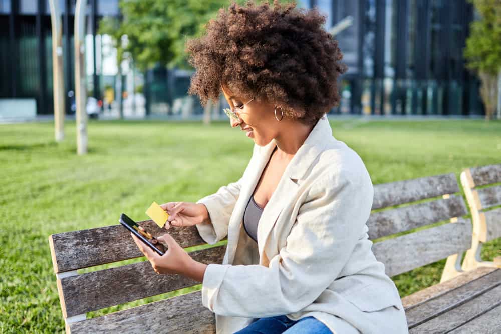 Young black female researching purple shampoo and DIY hair dyes using her cell phone while sitting on a park bench.
