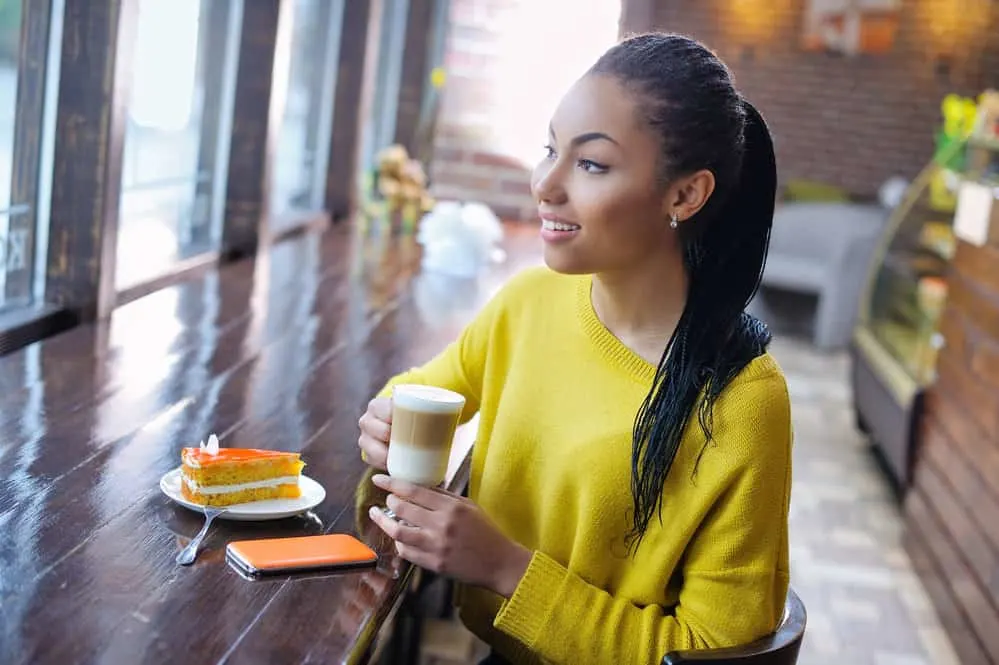 A young light-skinned black lady wearing micro locs and a yellow sweater while drinking a coffee in a donut shop.