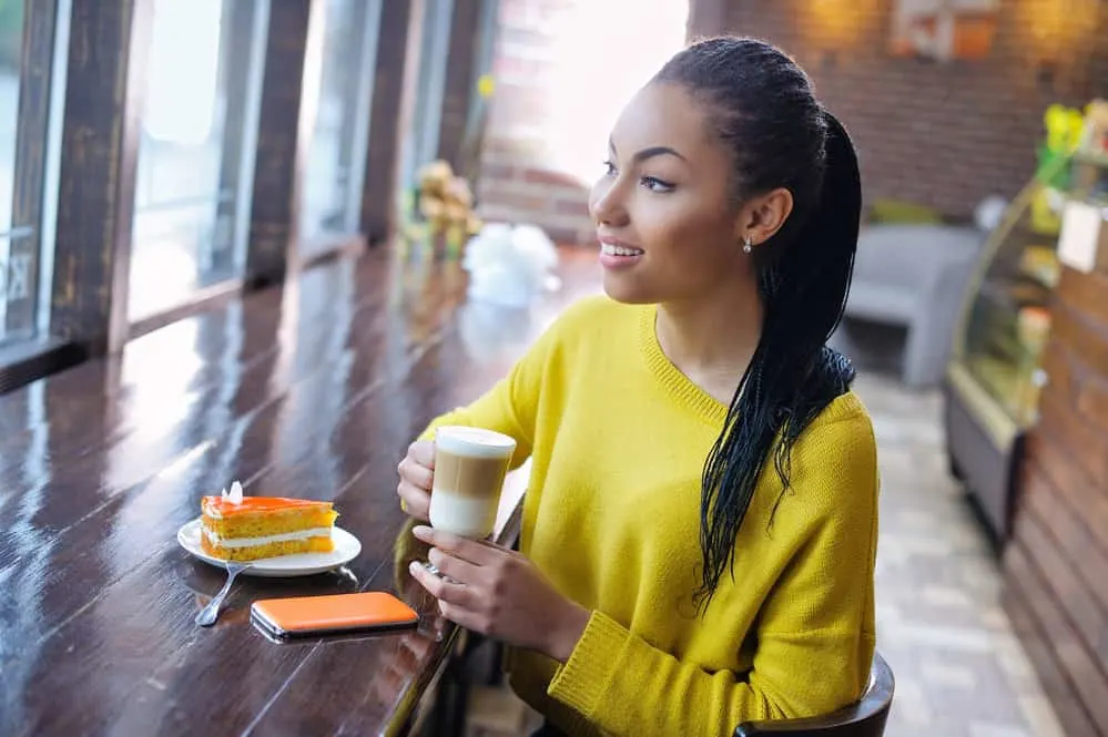 Microlocs: Styles, Installation, Cost, Products, and More 7 A young light-skinned black lady wearing micro locs and a yellow sweater while drinking a coffee in a donut shop.