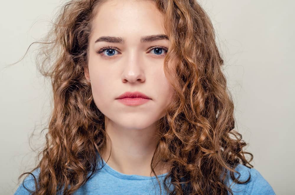 A white lady wearing a blue shirt with blue eyes has wavy textured hair strands created with styling mousse.