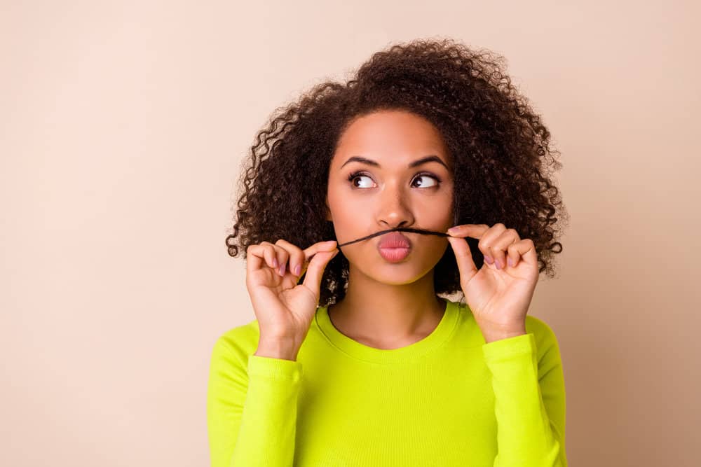A cute African American female holding a hair strand across her lip as a fake mustache wearing a green casual shirt.
