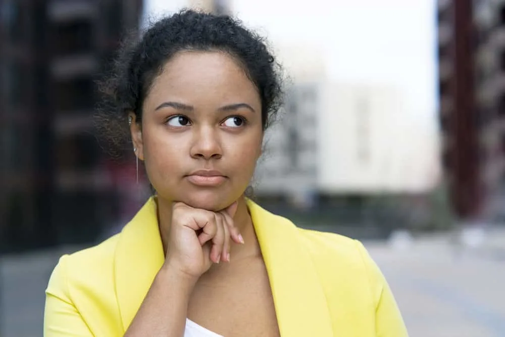 A female with a curly hair type wearing a yellow sports coat while pondering whether straightening treatments are safe.
