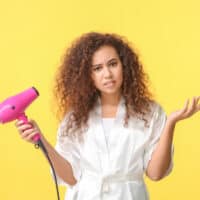 Beautiful African-American female with wet hair using a dryer on maximum heat on her thin hair strands.