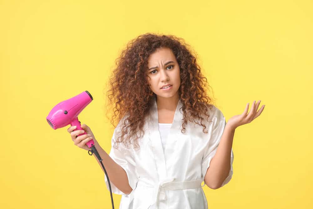 Beautiful African-American female with wet hair using a dryer on maximum heat on her thin hair strands.