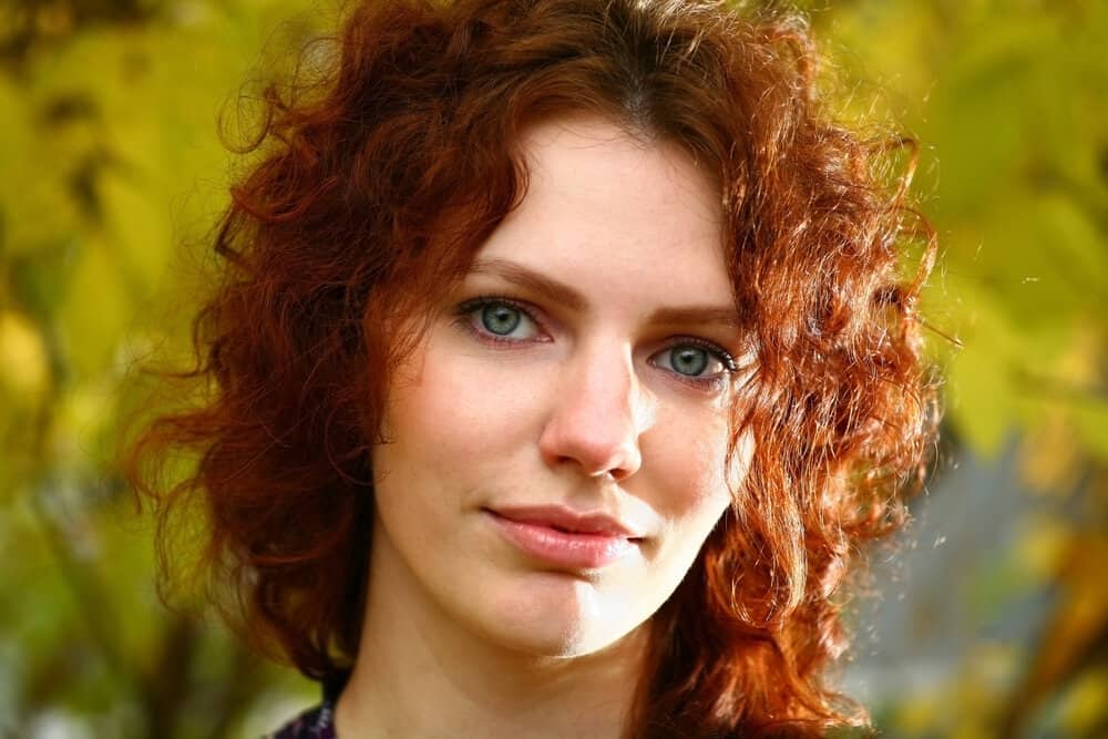 A joyful female with deep red shoulder-length hair has a type 3 curl pattern on her curly frizz-free natural curls.