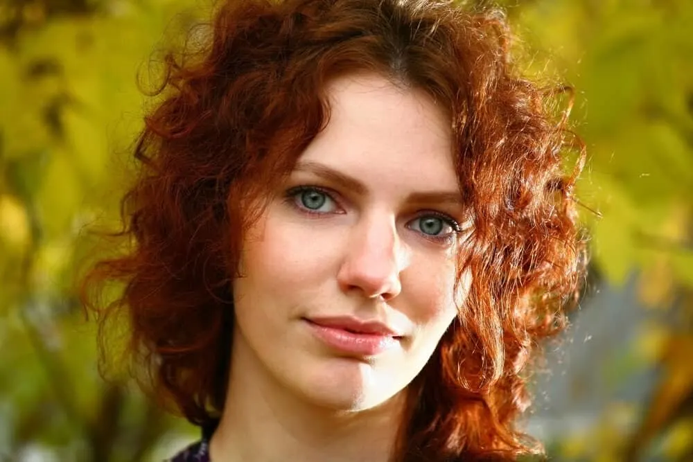A joyful female with deep red shoulder-length hair has a type 3 curl pattern on her curly frizz-free natural curls.