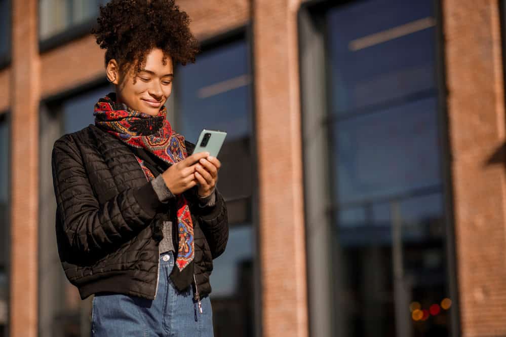 A female with a curly natural hair type is wearing blue jeans and a black jacket while texting a friend.