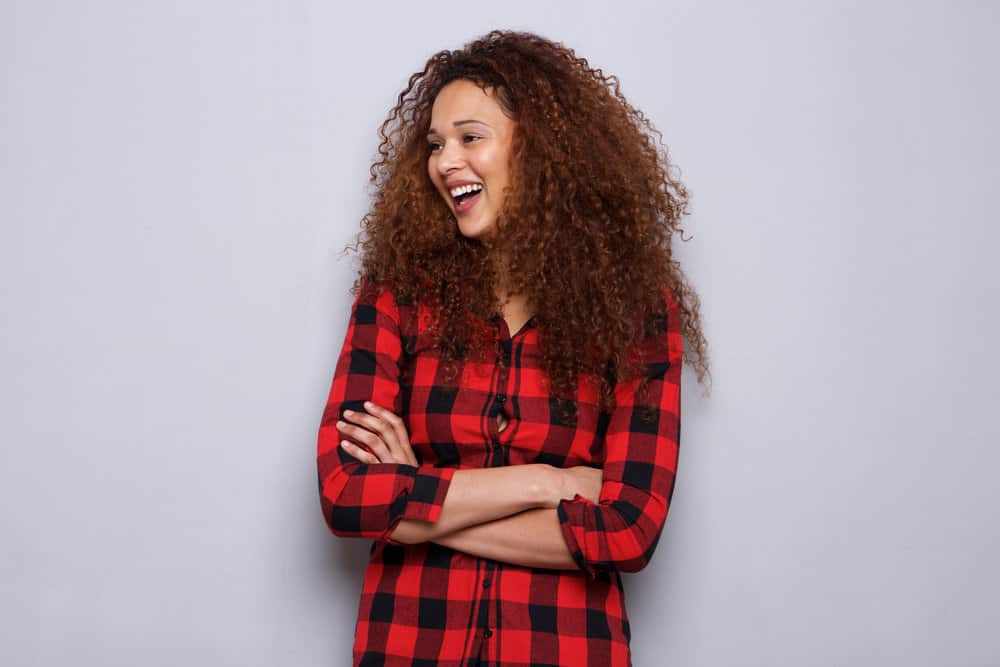 A young woman with natural curls dyed with demi-permanent hair color has her arms folded while laughing.
