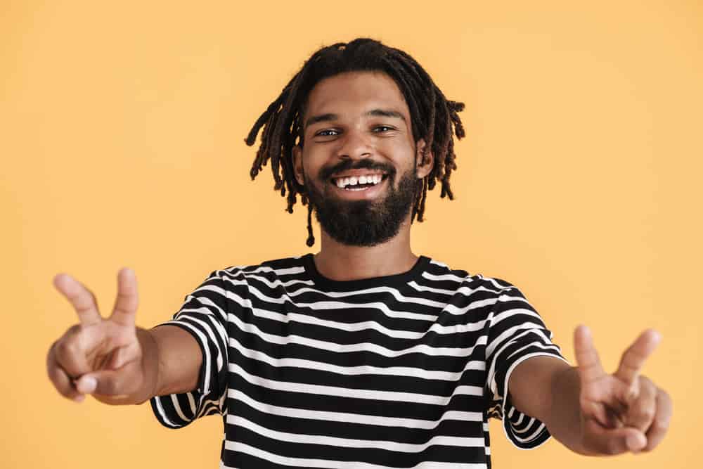 A young black guy with mature dreads and a few baby locs is making a peace sign with his hands.