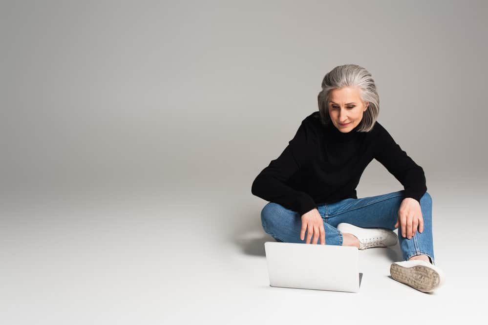 An older lady with naturally grey hair starting to grow in is researching silver hair and purple shampoo on her laptop.