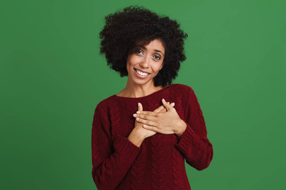 Black young woman smiling and holding hands on her chest isolated over green background