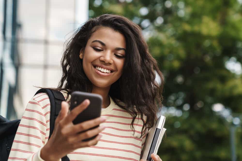 A cheerful young black female reading an article about how to flat iron hair on her mobile phone while outdoors.