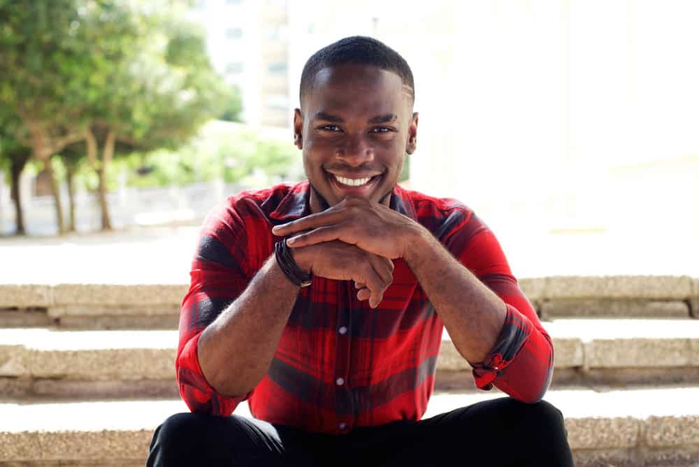 A dark skin young guy sitting outdoors on steps wearing a low-drop fade with a custom fade line on the left side.