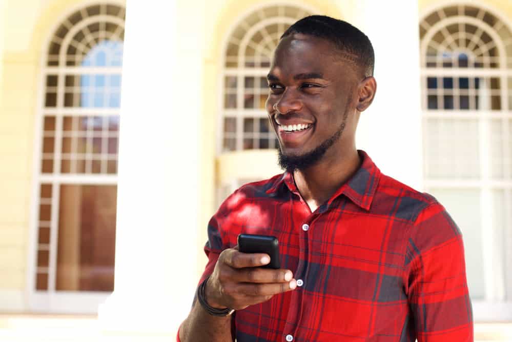 A dark skin black male with a short fade haircut on a mixture of type 4 hair types is standing outside using an iPhone.