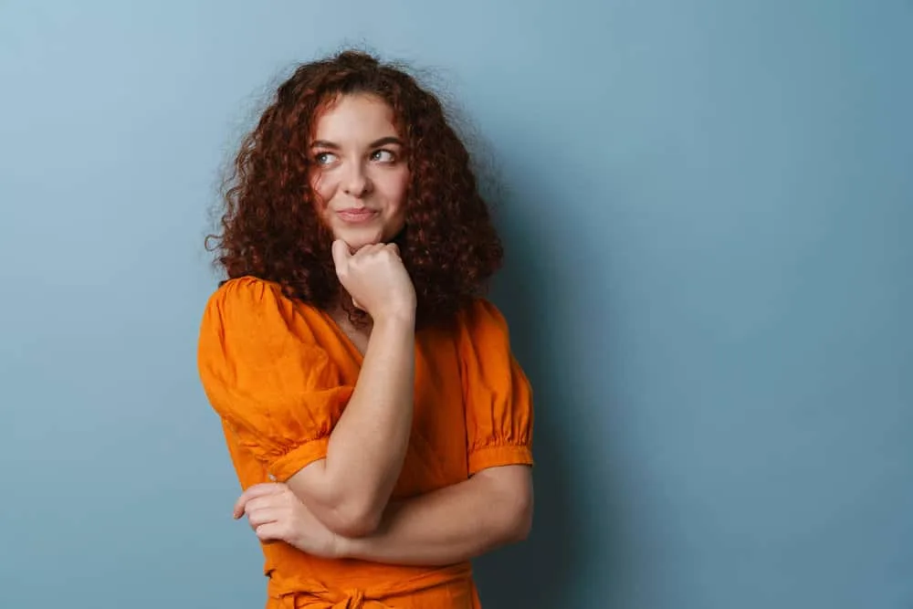 Get a Perm Out: How to Unperm Your Hair Naturally at Home 12 A beautiful lady with a subtle smile wearing an orange dress has her hair styled with a curling iron.