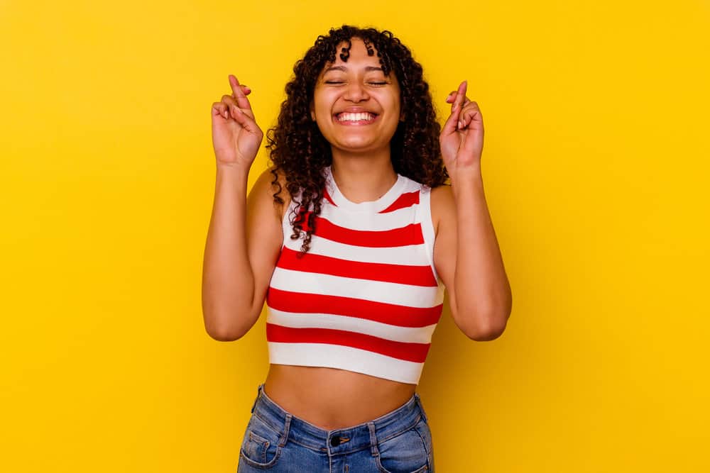 A young black female with kinky, curly hair strands is crossing her fingers, hoping that the Wave Nouveau perm works.