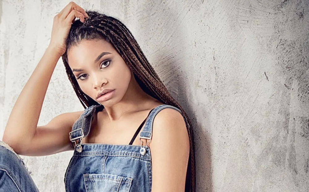 A fashionable female having her hair braided in a protective hairstyle is wearing blue jean overalls.