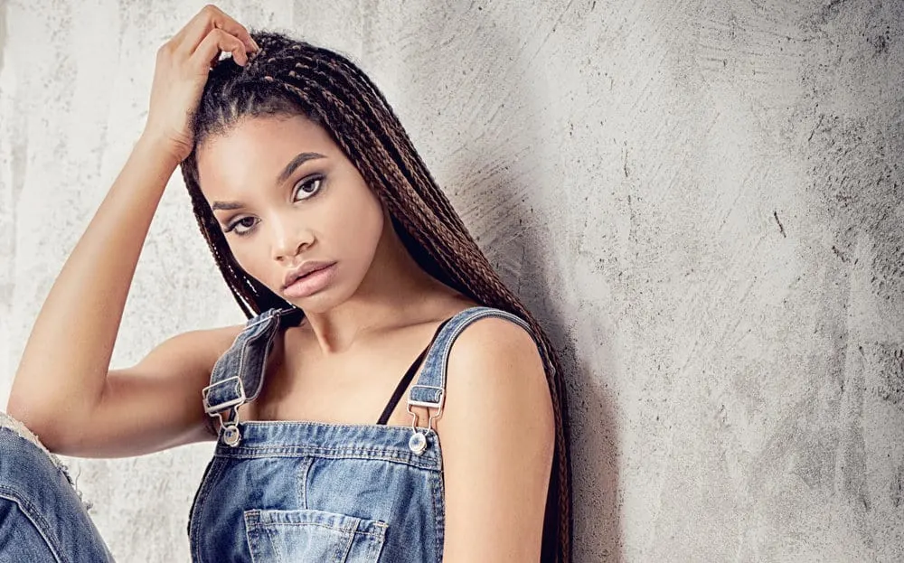 How Long Do Micro Braids Take, Last, Cost, and Other FAQs 3 A fashionable female having her hair braided in a protective hairstyle is wearing blue jean overalls.