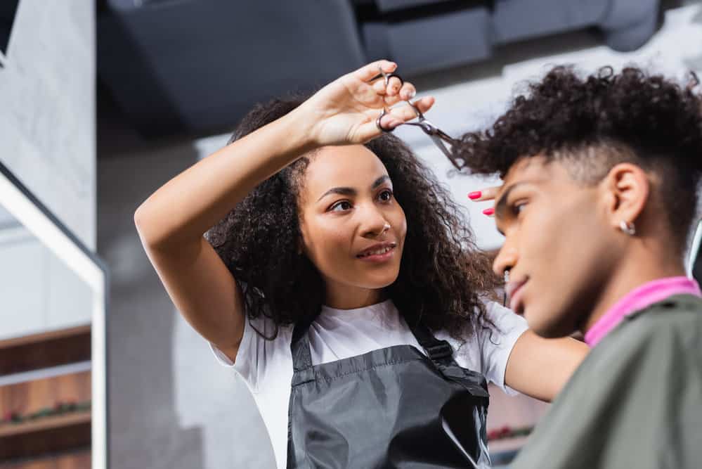 A black female hairdresser dried her client's wet hair and is now cutting his bangs using an average pair of shears.