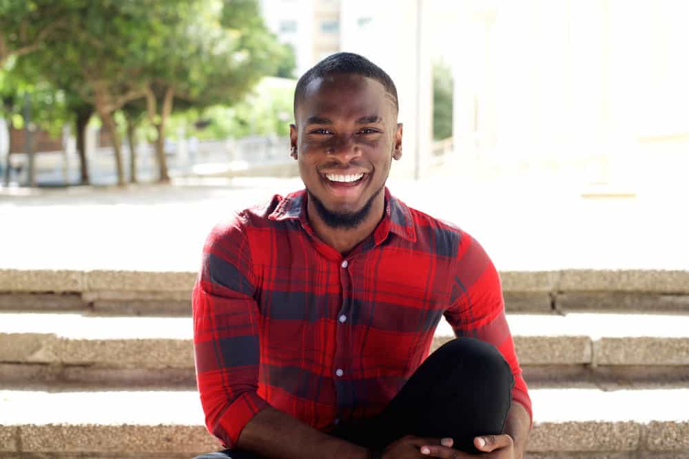 A man sitting outside and smiling while wearing a medium fade haircut on an oval face shape.