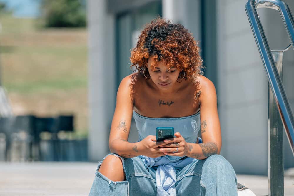 A stylish young female with dry hair researching how to use a deep conditioner for her hair type on her iPhone.