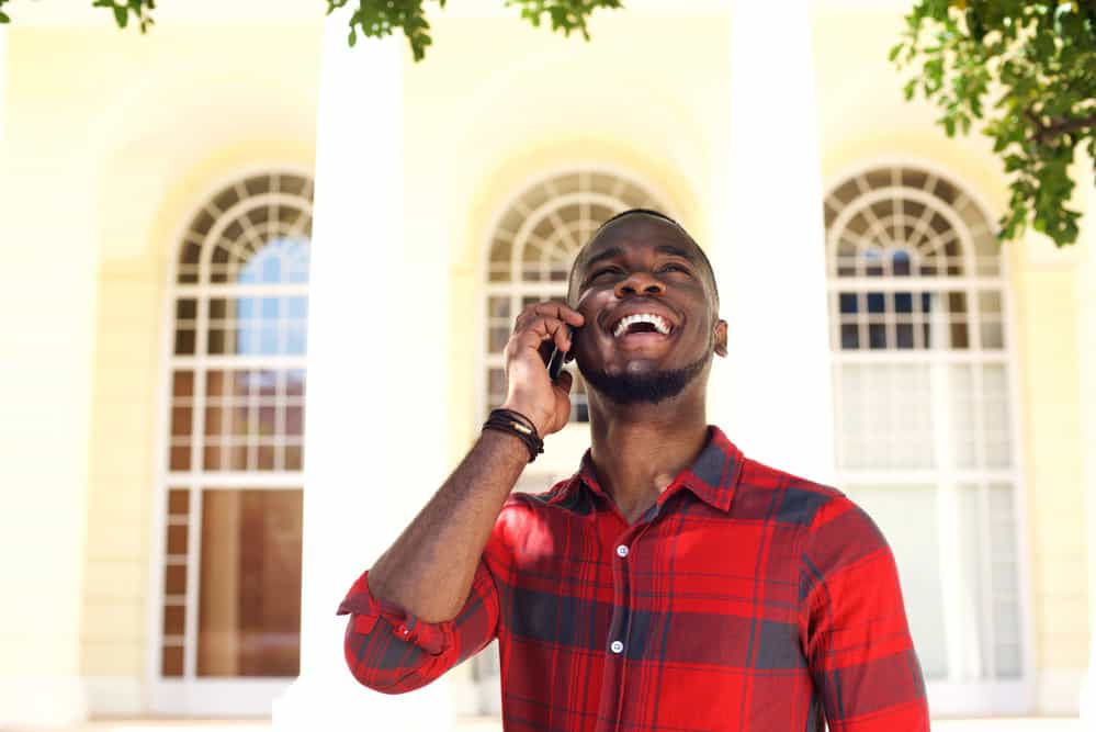 African American man with a bald drop fade and textured hair is standing outside talking on the phone with friends.