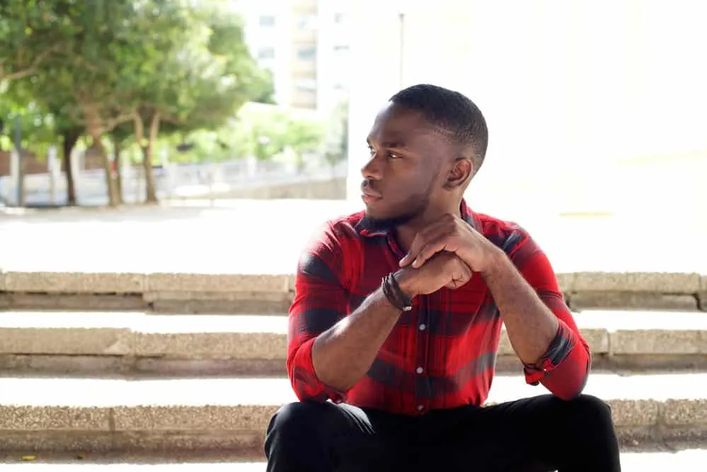 28 Different Types of Fades and Tapered Haircuts for Men 38 A calm young black male wearing a very short hair cut on his type 4 hair type is sitting outside relaxing.