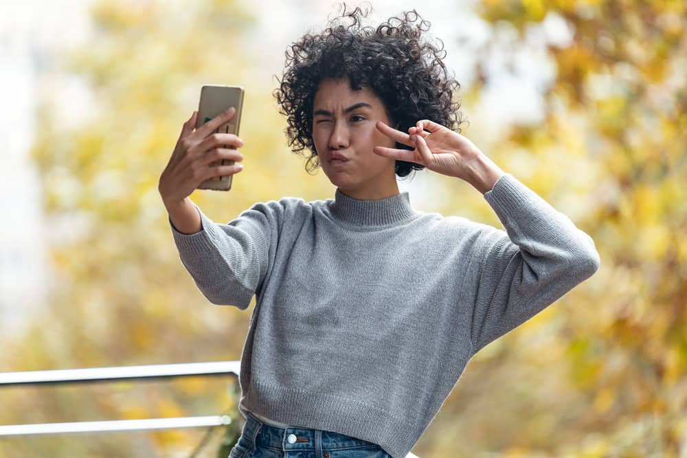 A beautiful woman with shiny natural curls takes a selfie photo while making a peace sign with her hand.