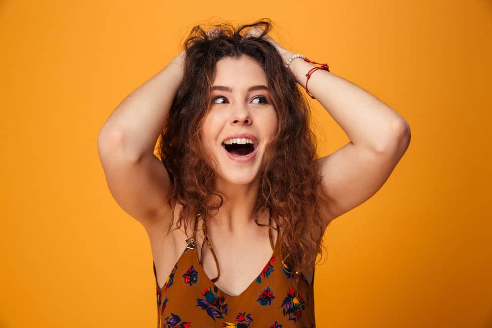 A female wearing a brown dress with red and green flowers is holding her messy hairstyle with both hands.